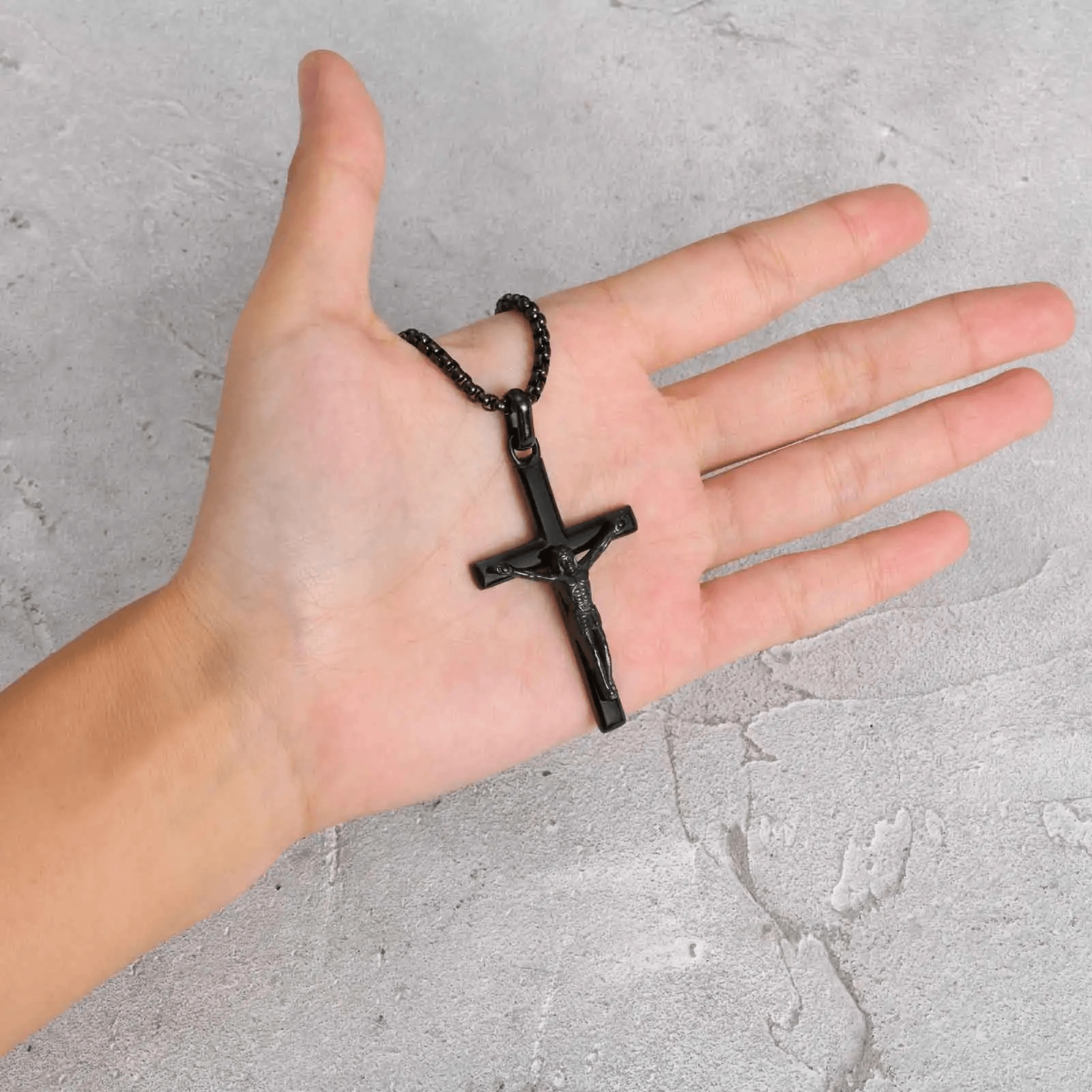 Hand holding a black cross necklace against a gray concrete background